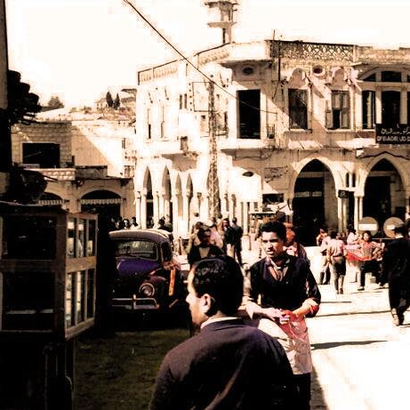 A lively street scene in front of a building with arched windows and a sign in Arabic and French. People are walking, and cars are parked nearby.