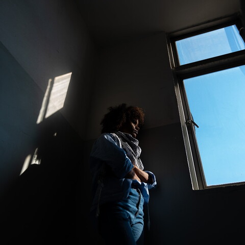 A photo taken from below; a woman stands by a window in a dark room. The sky outside is a clear blue. The sunlight hits her softly.