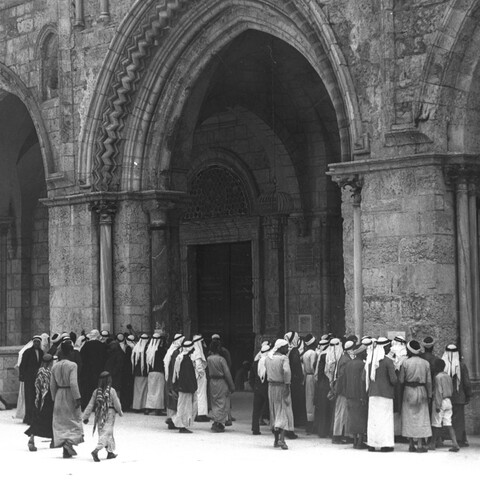 A black-and-white photograph of a group of men gathered outside the arched entrance of al-Aqsa Mosque in Jerusalem. 