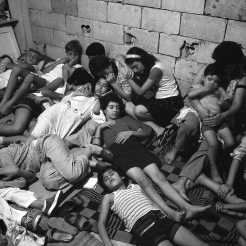 A black and white photo of a group of people leaning against one another, asleep on the floor, in what looks like a basement.