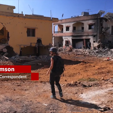 A woman stands amid rubble, center frame. Around her are fully demolished and semi-demolished buildings. She is wearing a helmet and flak jacket. To her left is a graphic that reads: “Lucy Williamson | BBC Middle East Correspondent”
