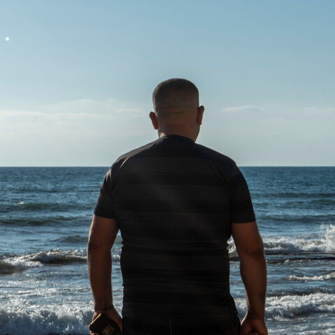 A man stands with his back to the camera and the sea before him. The sun is bright and the water is a deep blue. 