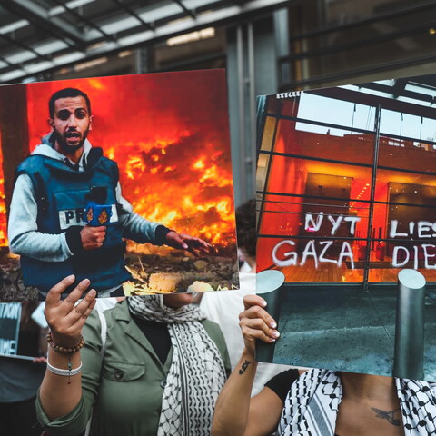 People hold up posters and signs at a protest. The poster in the center of the image is a photo of martyred Al Jazeera journalist Anas al-Sharif, killed by Israel on August 10, 2025. Next to it is a poster that reads: "NYT LIES, GAZA DIES"
