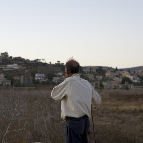 A man stands in a dry field looking toward a hillside town at dusk.