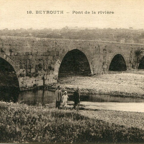An arched stone bridge crosses the Beirut River; mountains are visible in the background. Three people are walking by the river. 