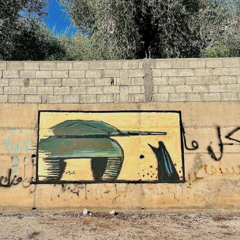 A mural of a green military tank is painted on a concrete wall with graffiti and a low brick barrier above; a woman is confronting the tank; trees and blue sky are visible in the background.