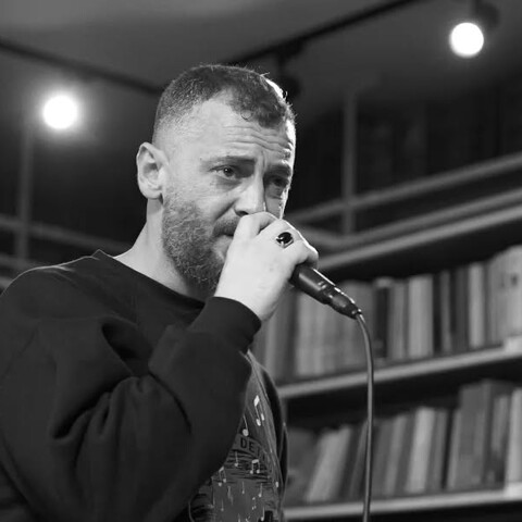 A bearded man passionately sings into a microphone, set against a backdrop of bookshelves. The black and white image conveys an intimate, artistic atmosphere.