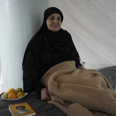 An older woman dressed in black sits beside a bed inside a tent-like shelter, holding a folded blanket. A small plate of fruit and a book rest on the bed next to her.
