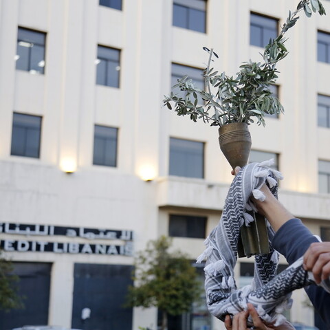 A hand holds remains of a missile filled with olive branches. A kuffiyeh is wrapped around the hand.
