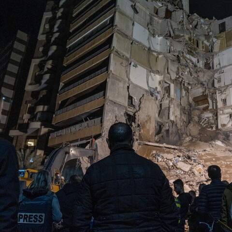 Crowd gathers at night before a partially collapsed building, with exposed interiors and debris. Somber scene with press and onlookers observing.