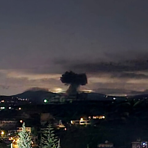 A dark plume of smoke rises into the night sky above a distant city, with lights from buildings visible in the foreground.