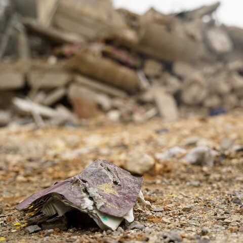 Tattered papers lie on rocky ground with a collapsed building in the background,
