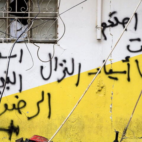 Graffiti in Arabic covers a white and yellow exterior wall beneath a barred window with exposed wires hanging across the scene.