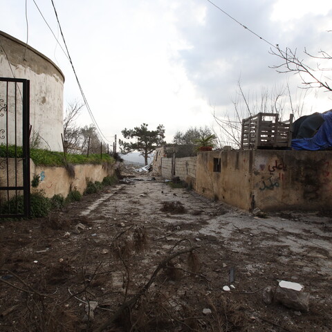 A narrow, muddy path between weathered concrete walls is flanked by a rusty gate and graffiti.