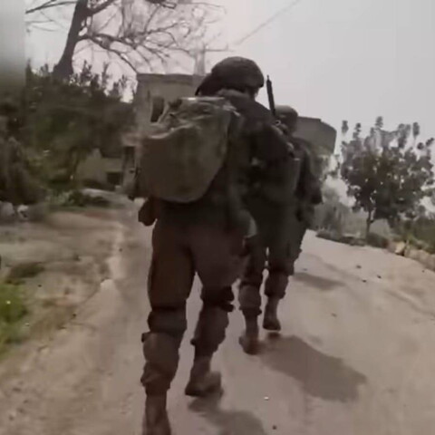 Two soldiers are running toward a house along a road lined with bushes and trees on both sides.
