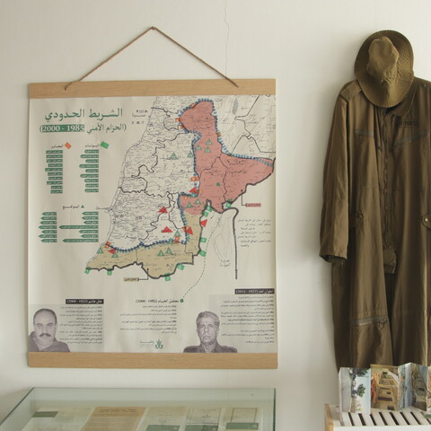 An exhibition wall featuring a large framed border map with Arabic text on the left, and a full-length olive-green military jumpsuit with a matching boonie hat hanging on the right.