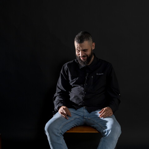 A man sits on a seat in front of a black background. He is bearded, wearing black, and looks down, laughing shyly.