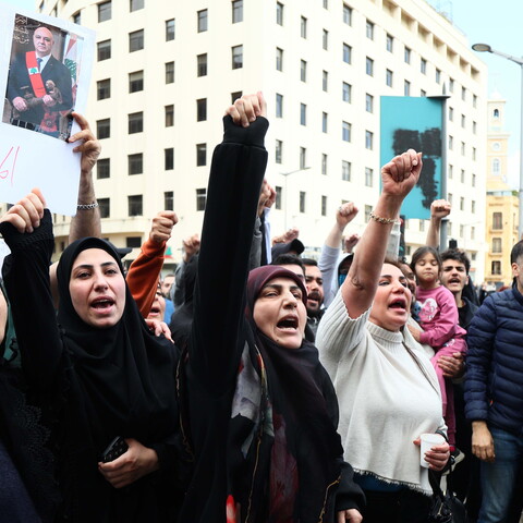 A crowd of people protest in a city street, raising their fists and shouting; one person holds a sign with Arabic writing and a photo of a man.