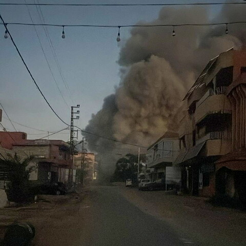 A street-level view in a residential neighborhood where a massive, towering plume of thick grey smoke rises high into the sky from directly behind the buildings on the right side of the road. A string of bare light bulbs hangs across the street under a dim sky.