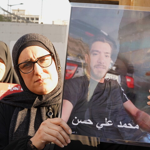 A woman in a black hijab and glasses holds up a large, laminated portrait of a missing man named Mohammad Ali Hassan during an outdoor demonstration. Other women stand closely behind her in solidarity.