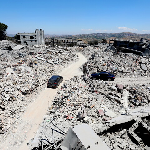 A high-angle panoramic view of massive structural destruction in Tebnine, South Lebanon. Mounds of shattered concrete and exposed rebar line a curved dirt road that has been cleared through the ruins. A dark gray hatchback and a black sedan are navigating the road amidst the completely flattened landscape.