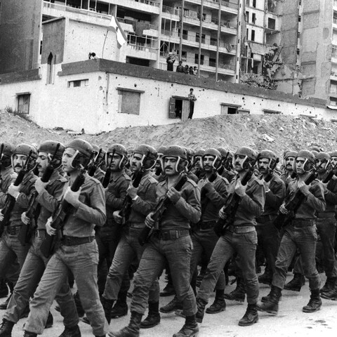 A black-and-white photograph of a large formation of Lebanese Army soldiers marching in tight unison during a drill. The soldiers are dressed in matching uniforms and combat boots, wearing distinctive helmets with integrated ear and chin protection, holding rifles diagonally across their chests. In the background, a war-torn urban landscape features large mounds of dirt and rubble in front of a heavily damaged, multi-story apartment complex and a smaller white building marked with bullet holes.