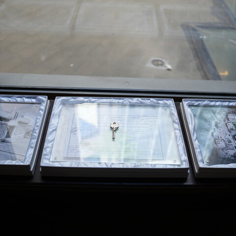 Three framed displays rest on a windowsill, each containing a key placed over a visual object: a photograph of a damaged house, a title deed, and an aerial image of a marked area. 