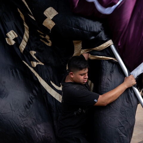 A young man wearing a black shirt strains as he hoists a large metal pole carrying heavy black and maroon religious banners trimmed with gold Arabic calligraphy during Ashoura commemorations.