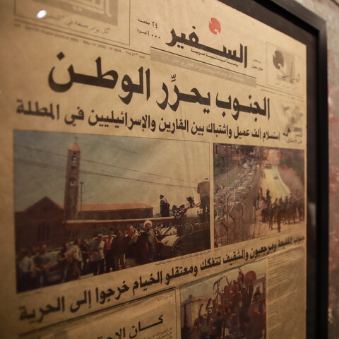 A framed Arabic newspaper with images of crowds, soldiers, and vehicles hangs on a textured wall next to a fire alarm.
