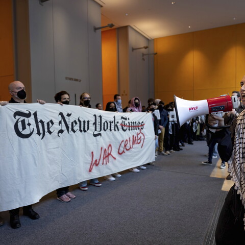Protesters at a demonstration inside The New York Times building, holding a banner accusing the publication of coverage related to war crimes, with one person speaking through a megaphone.