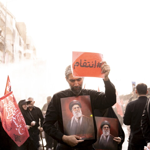 A man dressed in black walks through a crowd holding a framed portrait of Ayatollah Ali Khamenei against his chest while raising a red sign with the Arabic text "#انتقام" (#Revenge) in the air. Red flags and other mourners carrying portraits are visible in the background.