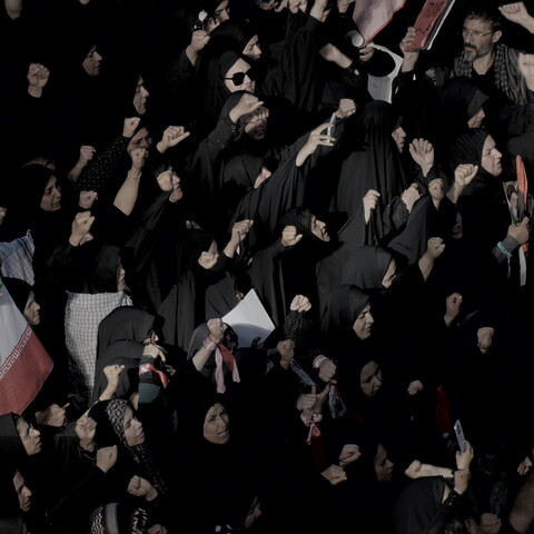 A high-angle view of a tightly packed crowd, largely composed of women in black chadors, raising their clenched fists together during a large gathering, with an Iranian flag visible among the group.