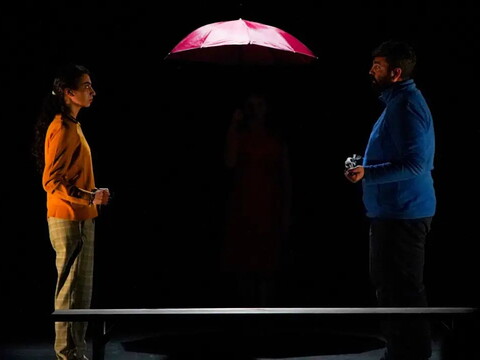 Two actors stand in the dark facing each other under a red umbrella.