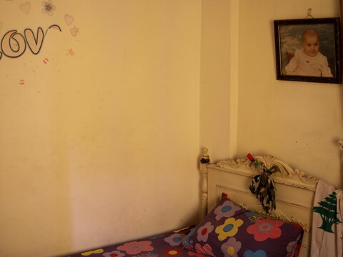 Picture of an empty purple bed with a flower pattern. A “LOVE” wall sticker is starting to peel off; on the bed is tied an army scarf holding up a plastic red rose.