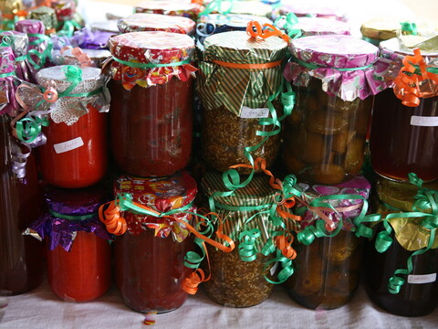 A variety of glass jars filled with preserved foods, such as jams and pickles, stacked on a table. The jars are decorated with colorful fabric covers and ribbons