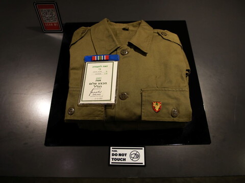 A folded olive-green military uniform displayed under glass, with an Israeli army badge, medal ribbon, and certificate dated July 8, 1983, placed on top.