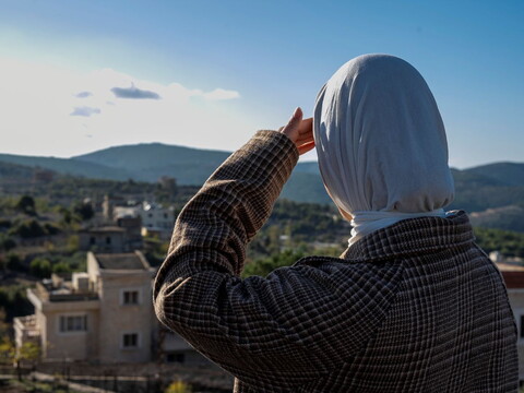 Hijabi woman sitting on a balcony overlooking rolling hills, with an Israeli military post visible on the highest mountain in the distance.