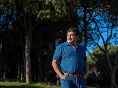 Man wearing a blue t-shirt and jeans, standing in a forest with trees visible in the background.