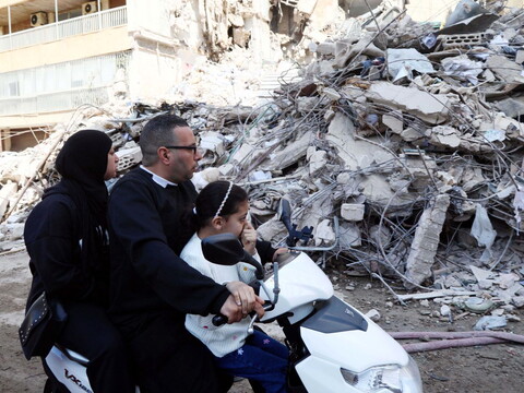 A man, woman, and child on a motorcycle pass by a large pile of rubble from a collapsed building in Beirut after an Israeli airstrike.