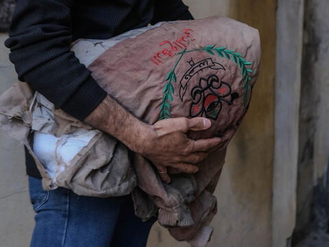 A person in a black sweater and jeans cradles an embroidered bundle, featuring green leaves and red symbols