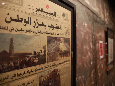A framed Arabic newspaper with images of crowds, soldiers, and vehicles hangs on a textured wall next to a fire alarm.