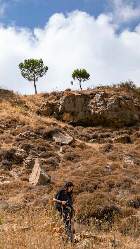 A wide shot showing a young woman dressed in black making her way through a foraging trail down a rocky path. Trees line up the top of the hill and stand tall against a cloudy blue sky.