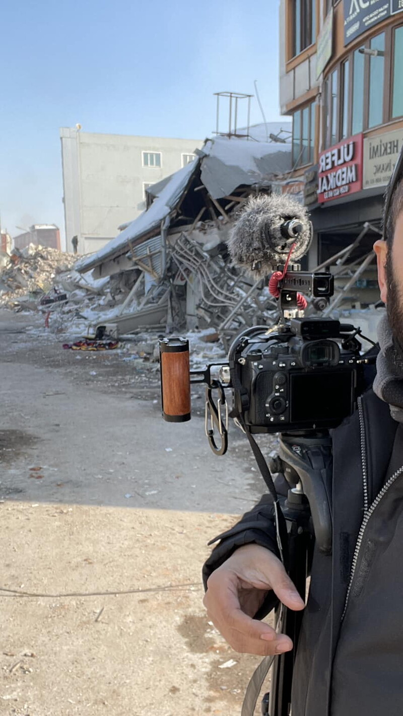 A self-portrait of journalist Issam Abdallah in the foreground, carrying his DSLR camera and microphone, to the background of destroyed buildings. Clear winter day.