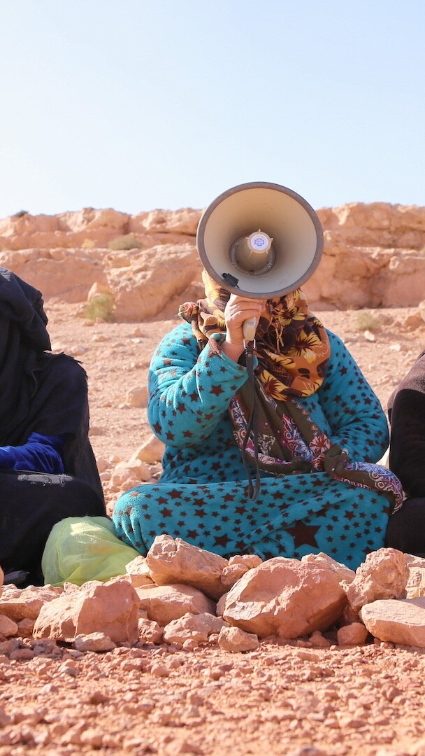 مجموعة من ست نساء أمازيغيات يجلسن على الأرض كتفًا لكتف. جميعهن لديهن شعر ووجوه مغطاة جزئيا؛ واحدة منهن تتحدث في مكبر الصوت.