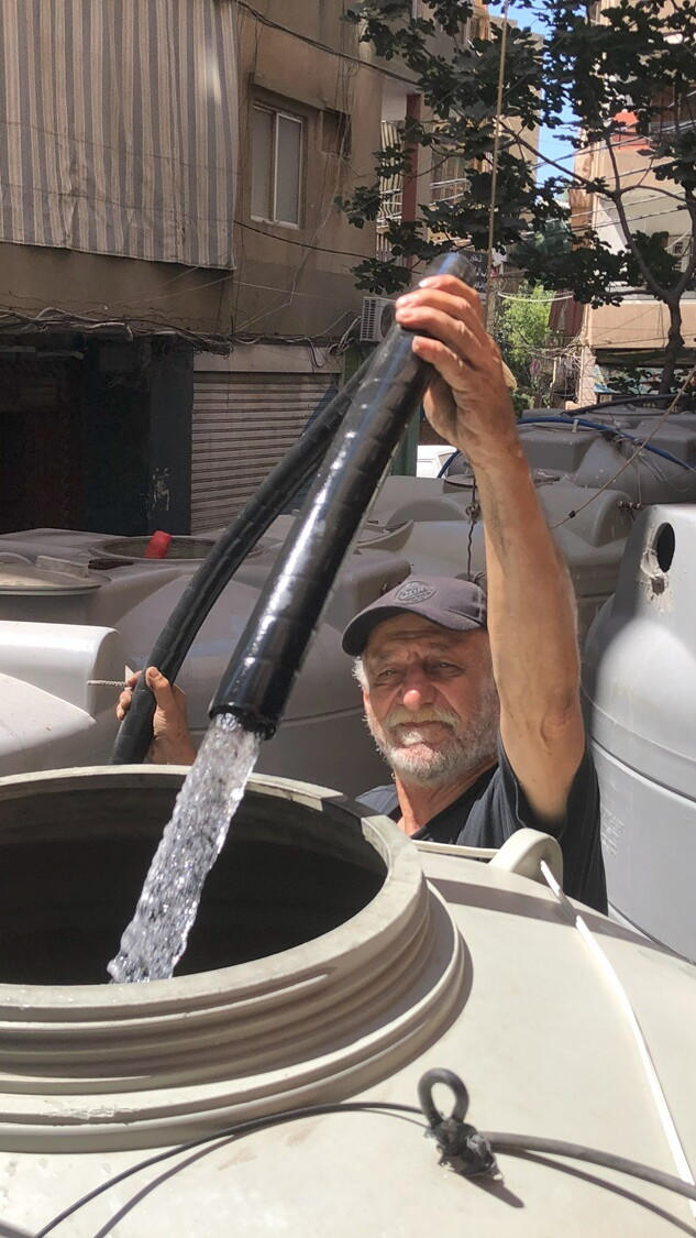 A man in a cap with a white mustache points a thick black hose into a plastic grey water container, and water flows from the hose into the container. There are other such containers around him.
