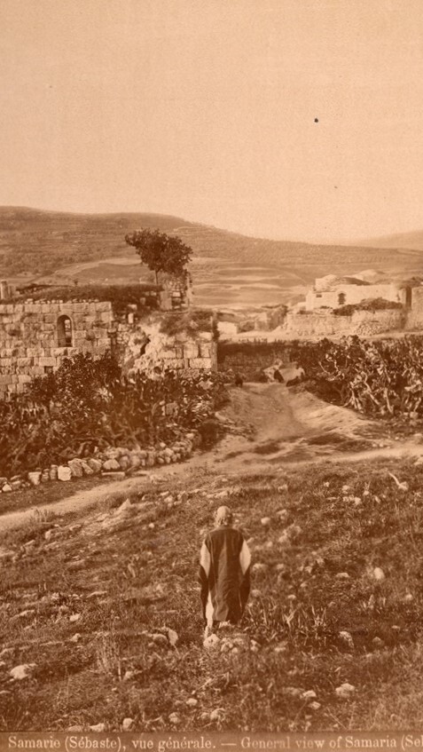 Sepia-toned photograph of ancient stone ruins and surrounding landscape in Samaria (Sebaste), with a person sitting in the foreground. Hills and buildings are visible in the background.