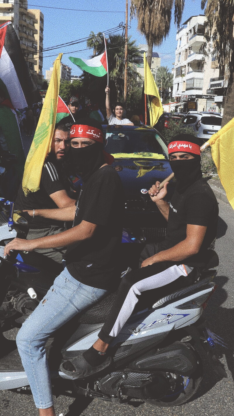Three male protestors on two motorcycles are carrying the yellow flag of Hezbollah. The Palestinian flag waves in the background.