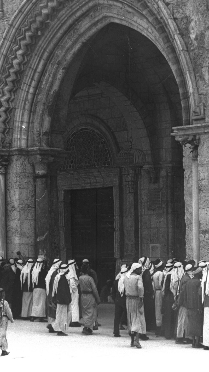 A black-and-white photograph of a group of men gathered outside the arched entrance of al-Aqsa Mosque in Jerusalem. 