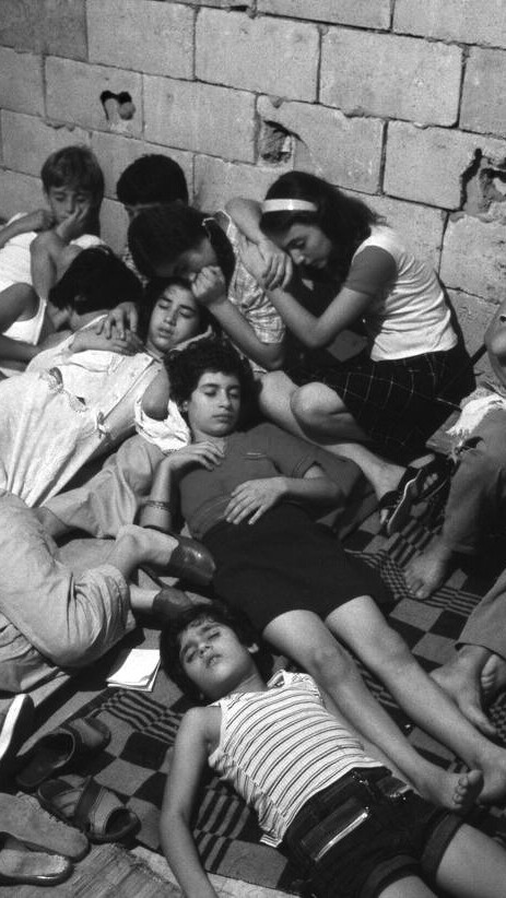 A black and white photo of a group of people leaning against one another, asleep on the floor, in what looks like a basement.