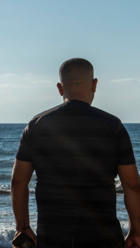 A man stands with his back to the camera and the sea before him. The sun is bright and the water is a deep blue. 